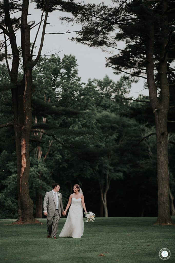 bride and groom walk hand in hand across the golf course under the trees at deerfield golf club in delaware