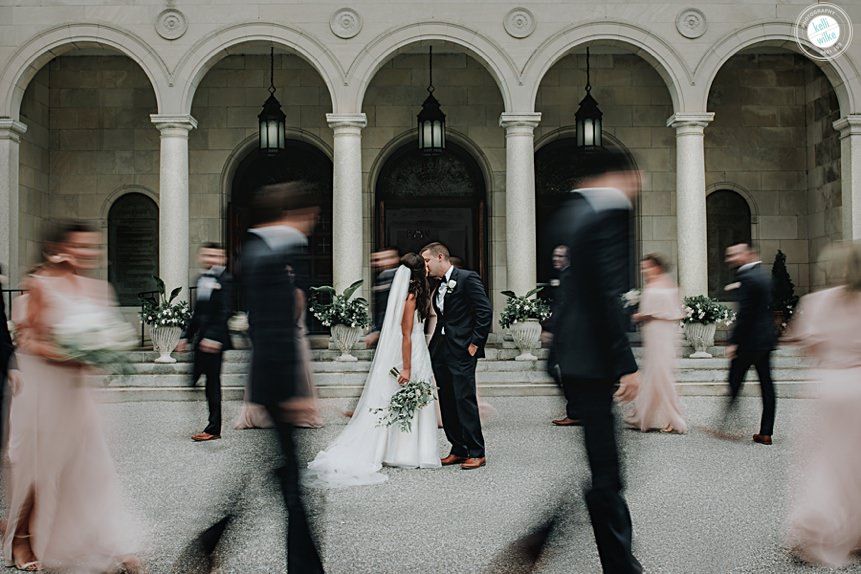 bride and groom standing still while the bridal party is in motion walking around the couple in front of a church in wilmington delaware photo was awarded masters of wedding photography award 