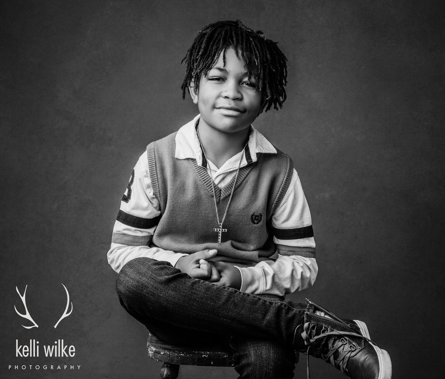 A black and white picture of a young boy sitting on a stool with his legs crossed, hands clutched, smirking at the camera. Captured by Kelli Wilke Photography. 