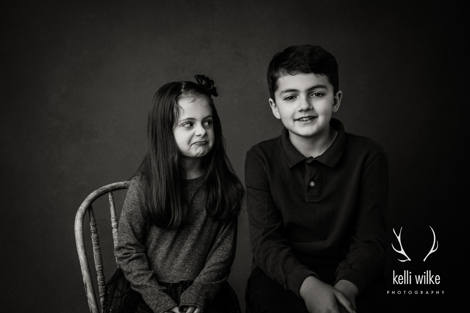 A black and white picture of a little girl making a silly face at her bigger brother as he smiles at the camera. 