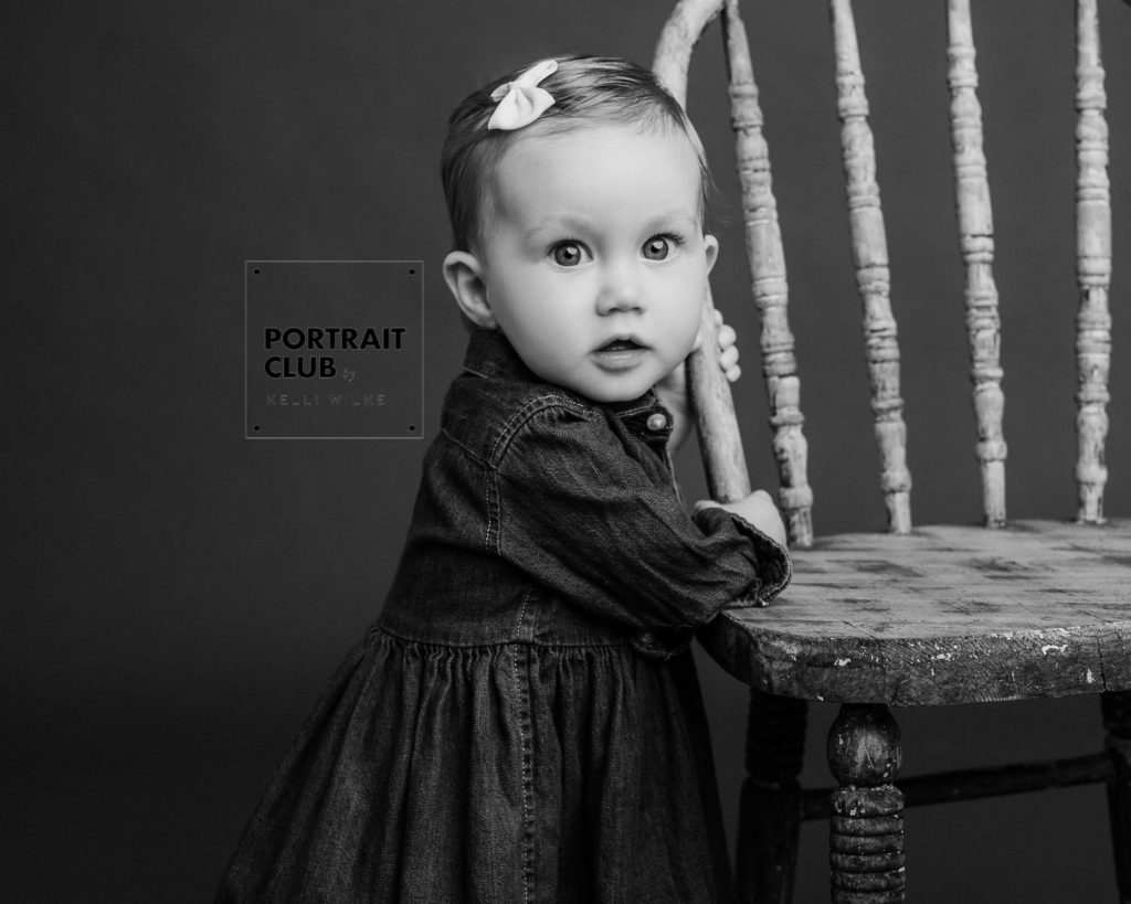 A candid black and white picture of a baby girl with a bow in her hair smiling and looking at the camera during a studio portrait session with Portrait Club by Kelli Wilke in Wilmington, Delaware. 