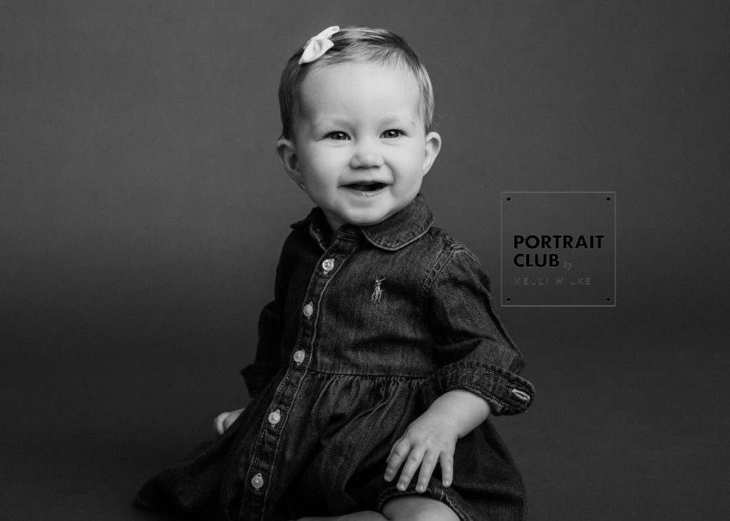 A candid black and white picture of a baby girl with a bow in her hair smiling and looking at the camera during a studio portrait session with Portrait Club by Kelli Wilke in Wilmington, Delaware. 