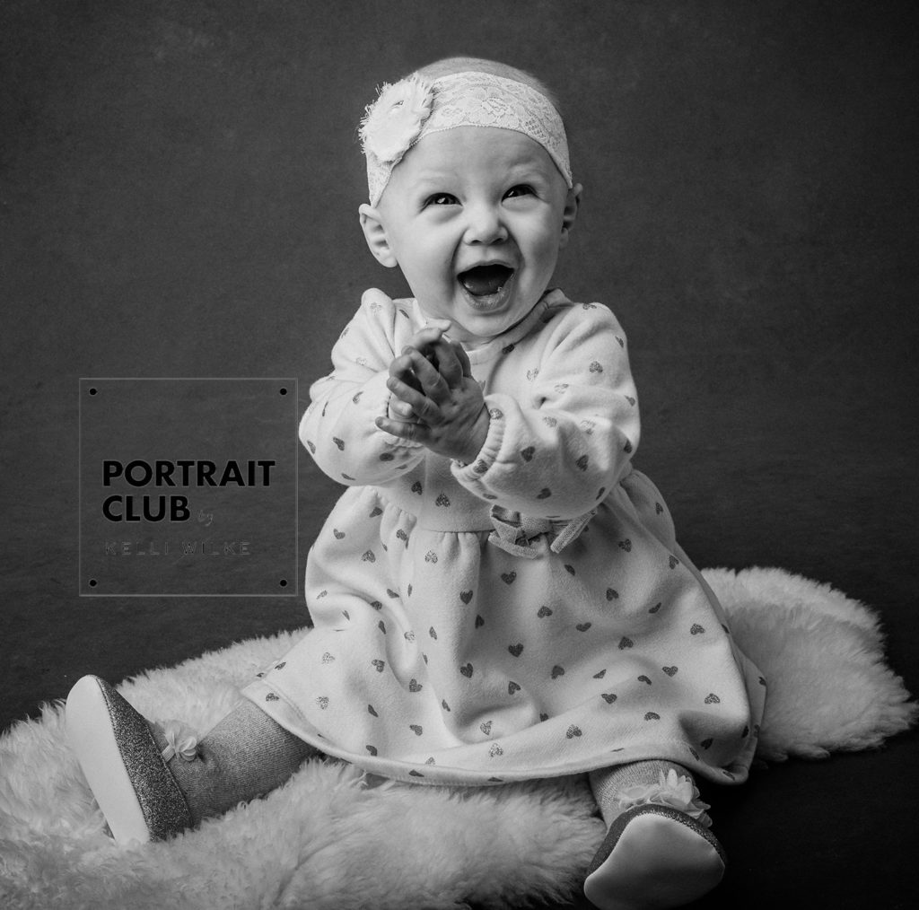 A candid black and white picture of a baby girl with a bow in her hair smiling and looking at the camera during a studio portrait session with Portrait Club by Kelli Wilke in Wilmington, Delaware. 