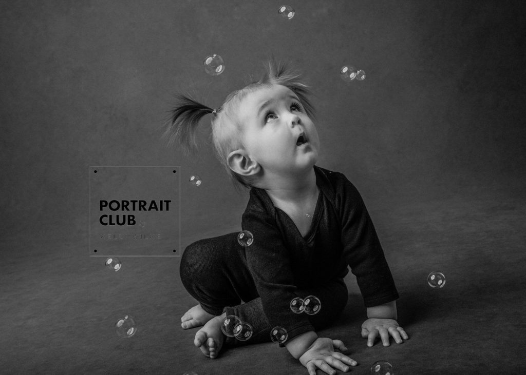 A candid picture of a little girl in pigtails looking at bubbles floating around her head in amazement during a studio portrait session with Portrait Club by Kelli Wilke Photography in Wilmington, Delaware. 